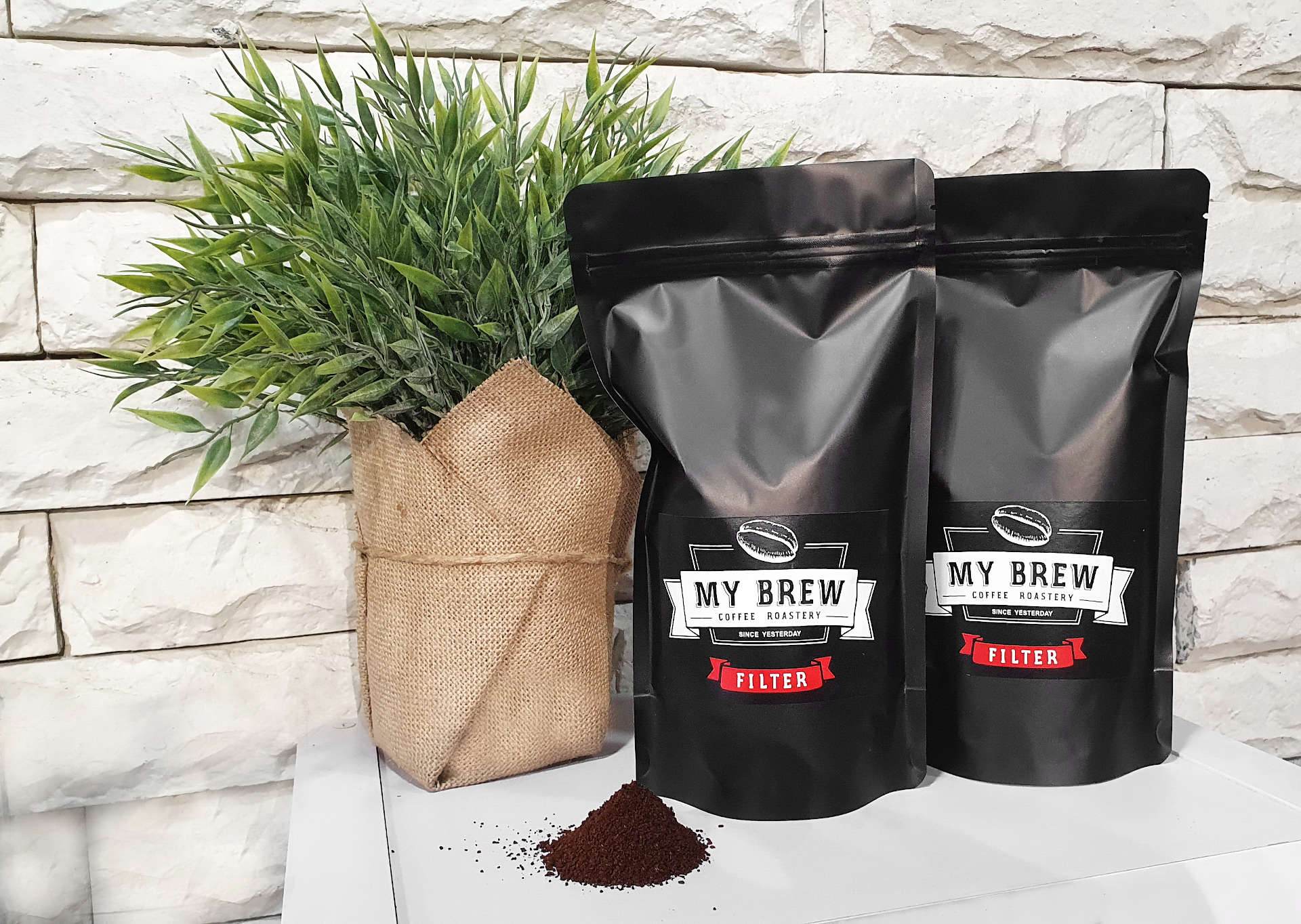 Two matte-black bags of My Brew Filter coffee with red labels beside a pile of ground coffee